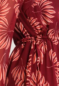 Robe rouge à fleurs avec manches longues, présentant une taille ajustée, un détail noué et un tissu doux et léger à texture plissée.