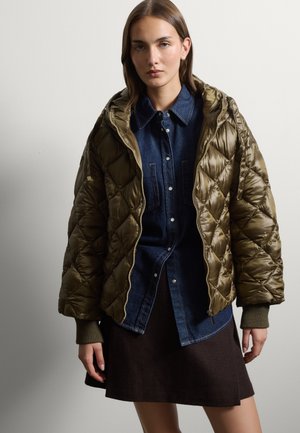Young woman wearing an olive green quilted jacket over a dark blue denim shirt and a dark brown skirt, standing against a plain gray background.