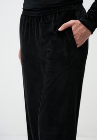 Pantaloni in velluto nero con una consistenza morbida, dotati di una cintura elastica e tasche laterali. Il tessuto ha una leggera lucentezza.