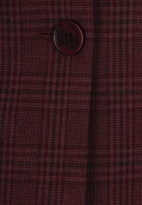 Tessuto a quadri bordeaux con una trama texturizzata. Presenta un bottone rotondo di colore scuro con la scritta "PATRIZIA PEPE" impressa.