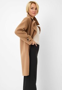 Camel-farbenen Mantel aus weichem Stoff, mit einem breiten Kragen, langen Ärmeln mit Knopfdetails und einem knielangen Design. Getragen über einem gerippten crèmefarbenen Oberteil.