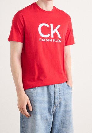 Mann trägt rotes T-Shirt mit kurzen Ärmeln und großem weißem "CK Calvin Klein"-Logo, kombiniert mit hellblauen Jeans, Hände locker.