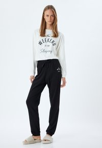 Camisa blanca de manga larga con texto negro, combinada con pantalones negros con cordón. El modelo lleva sandalias de peluche beige. Diseño simple y casual.