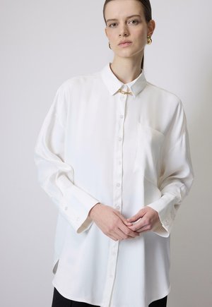 Kobieta z włosami zaczesanymi do tyłu, ubrana w oversize'ową białą koszulę na guziki, złote kolczyki koła, zapinająca mankiety koszuli na jednolitym tle.