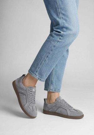 Persoon die lichtblauwe jeans en grijze suède sneakers met bruine zolen draagt, staand met één voet iets opgelicht.