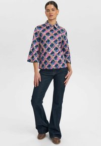 Camicia a maniche lunghe colorata con un motivo geometrico nei colori rosa, blu e beige; maniche a tre quarti; abbinata a jeans a vita alta blu scuro.
