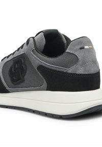 Zapatilla deportiva negra y gris con parte superior de malla y ante, suela acolchada y logotipo destacado en el lateral. Diseño minimalista con detalles texturizados.