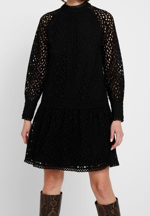 Schwarzes, durchlöchertes Kleid mit langen Ärmeln, hohem Hals und weit ausgestellt Rock. Mit kreisförmigen Ausschnitten, Spitzendetails und einer strukturierten Oberfläche.