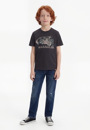 Αγόρι με κόκκινα μαλλιά που φοράει ένα μαύρο γραφικό T-shirt Wrangler, ξεθωριασμένα μπλε τζιν και μαύρα και άσπρα αθλητικά παπούτσια, στέκεται μπροστά σε λευκό φόντο.