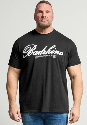 Muskelmann mit kurzen Haaren und Bart, der ein schwarzes "BadRhino"-T-Shirt und blaue Jeans trägt und vor einem schlichten hellen Hintergrund steht.