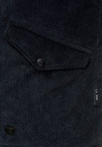 Nahaufnahme eines schwarzen Cordstoffs mit einer Klappentasche, die mit einem schwarzen Druckknopf gesichert ist, und kleinen schwarzen Markentags an der Seite und in der Ecke.