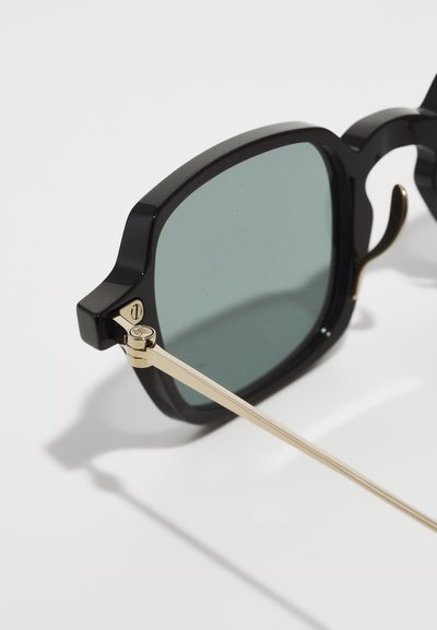 Schwarze rechteckige Sonnenbrille mit grünen Gläsern, goldenen Metallbügeln und einem ausgeprägten dicken Rahmen. Auffälliges Gelenkdetail in der Nähe der Gläser.