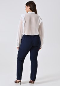 Donna con lunghi capelli scuri, che indossa una camicetta bianca trasparente con ruches, pantaloni blu navy e scarpe con tacco alte marroni, in piedi di spalle.