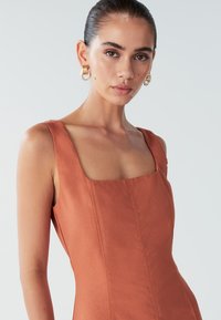 Šaty v rezavé barvě se čtvercovým výstřihem, širokými ramínky, přiléhavým střihem a jemnými švy. Modelka má na sobě zlaté kruhové náušnice. Hladká textilie.