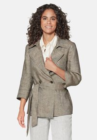 Lichtbruine, gestructureerde blazer met dubbelknopsluiting, brede revers en een strik taille, gecombineerd met een crèmekleurig shirt en lichte broek.