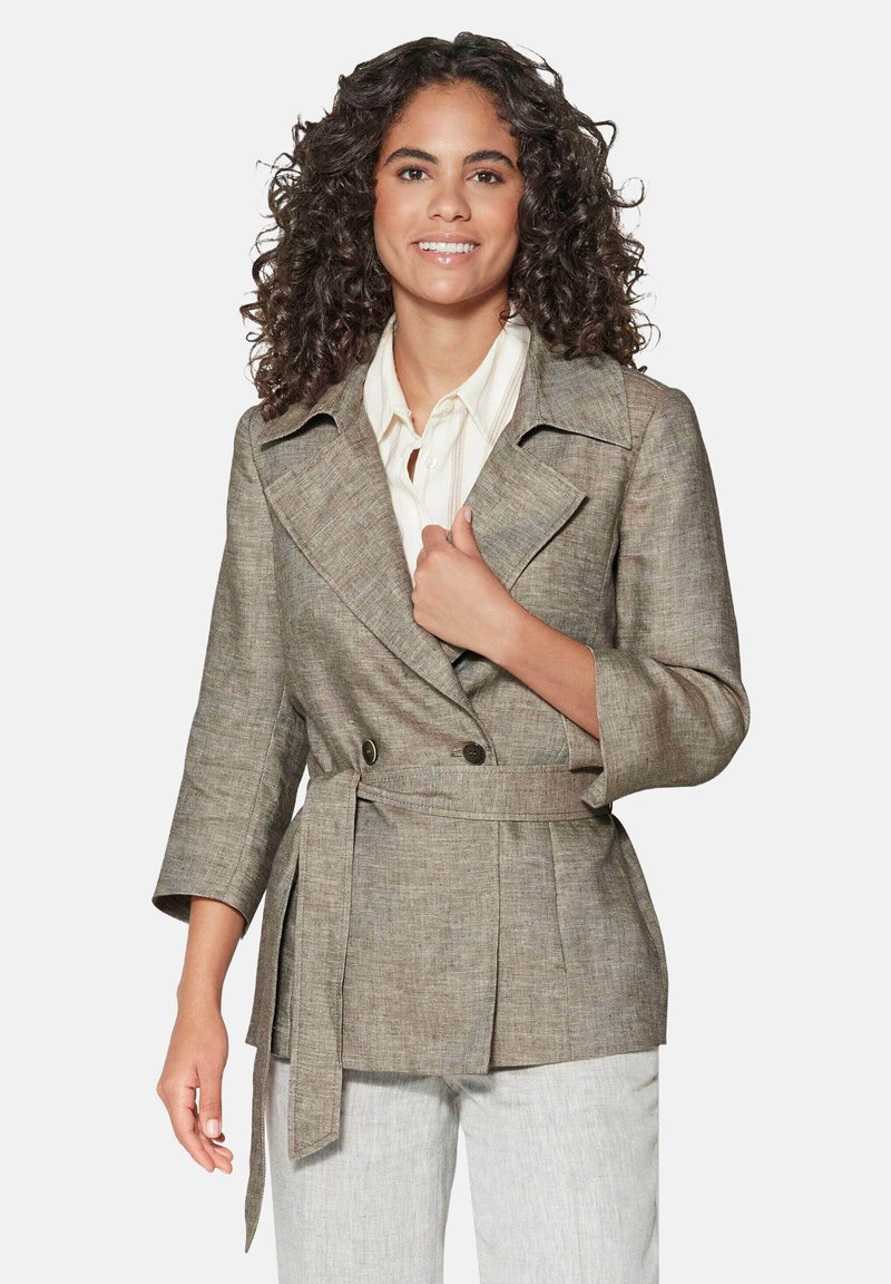 Lichtbruine, gestructureerde blazer met dubbelknopsluiting, brede revers en een strik taille, gecombineerd met een crèmekleurig shirt en lichte broek.