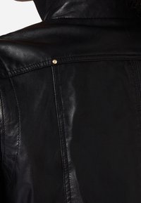 Černá kožená bunda s hladkou texturou, viditelným šitím a malým zlatým drukem na švu ramene.