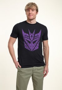 Zwarte T-shirt met een opvallend paarse grafische ontwerpen die lijken op een gestileerd logo, gemaakt van katoen of een katoenmix.