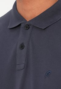 Mørkeblå polo skjorte lavet af tekstureret stof, med krave, tre sorte knapper og et lille broderet logo på brystet.