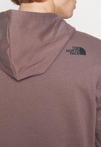 Sudadera con capucha de suave tela malva. Presenta una textura acanalada y un logo negro en la parte superior de la espalda. Corte estándar con capucha forrada.