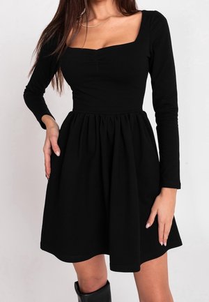 Femme portant une robe noire à manches longues, longueur genou, avec un décolleté carré et une taille froncée, associée à des bottes noires montantes jusqu'aux genoux.