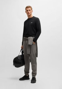 Cămașă neagră cu mâneci lungi și logo; pantaloni de trening gri cu fermoare pe lateral; purtând o geantă de sport neagră; adidași negri; stil sportiv casual.