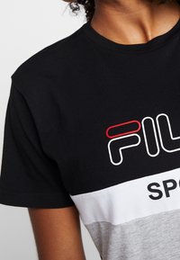 Detailní záběr na osobu, která má na sobě černé, bílé a šedé tričko FILA Sport s krátkým rukávem a viditelné kudrnaté vlasy.