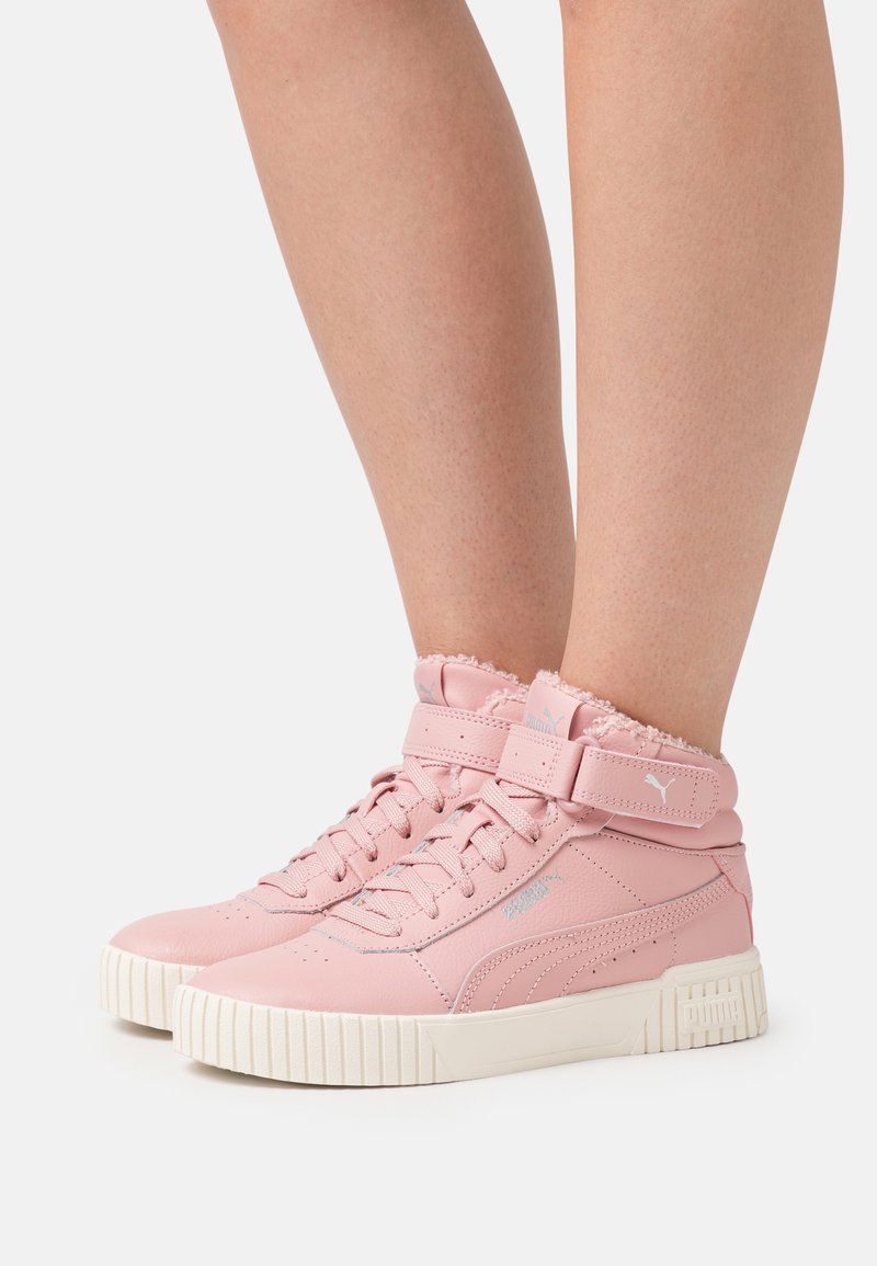 Jambes portant des baskets montantes roses avec des semelles blanches et des brides à la cheville sur un fond clair uni.