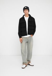 Zwarte ritsvest met grote voorklauwen, gedragen over een wit T-shirt. Lichtblauwe spijkerbroek en witte sneakers maken de outfit compleet. Zwarte beanie op het hoofd.
