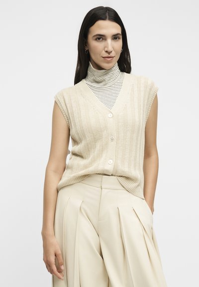 Γυναίκα με μακριά σκούρα μαλλιά φοράει κρεμ αμάνικη ζακέτα πάνω από ριγέ μπλούζα με ψηλό λαιμό και μπεζ πιετέ παντελόνια, στέκεται μπροστά σε λευκό φόντο.