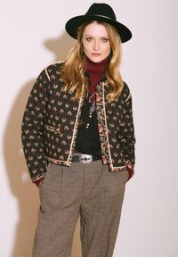 Veste matelassée noire à motifs floraux rouges et bordure beige, associée à un pantalon à carreaux gris et un col roulé bordeaux, avec une ceinture argentée.