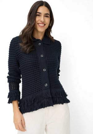 Lächelnde Frau mit dunklem, welligem Haar, die eine marineblaue, strukturierte, geknöpfte Strickjacke mit Fransen und weiße Hosen trägt, vor einem weißen Hintergrund stehend.