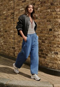 Jachetă bomber neagră peste un tricou henley cu dungi și pantaloni largi bleu deschis. Adidași albi completează ținuta casual în fața unui perete de cărămidă.