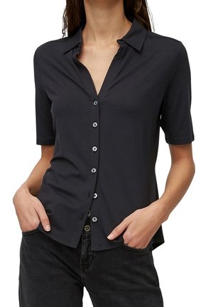 Vrouw draagt een nauwsluitend zwart overhemd met korte mouwen en kraag, handen in de zakken, gecombineerd met een zwarte spijkerbroek tegen een witte achtergrond.