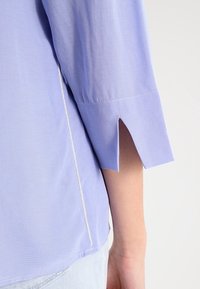 Blouse violet clair avec des manches trois-quarts, présentant des fentes sur les côtés et un subtil détail de couture décoratif le long de l'ourlet. Texture de tissu lisse.