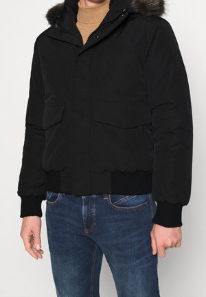 Mężczyzna w czarnej ocieplanej kurtce z kapturem obszytym futrem, beżowym golfie i niebieskich dżinsach, stojący na białym tle.