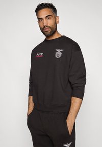 Fekete, rövid ujjú pulóver, bordázott mandzsettával és szegéllyel. Fehér és vörös hímzéssel rendelkezik, beleértve egy logót és szöveget az elülső részen.