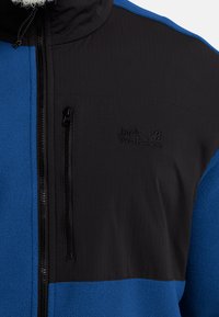 Veste en polaire avec une section inférieure bleue et un panneau supérieur noir. Dotée d'une poche poitrine zippée et d'un logo brodé. Tissu texturé.