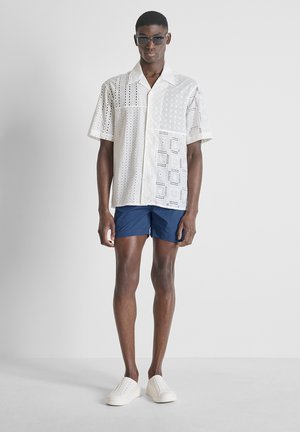 Man draagt een wit kortemouwhemd met patroon, marineblauwe korte broek, witte instapschoenen en een donkere zonnebril, staand tegen een effen achtergrond.