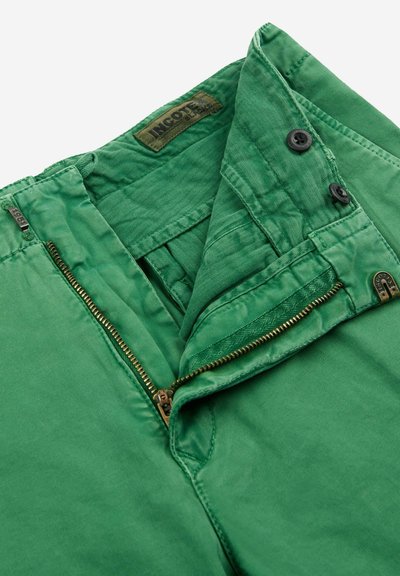 Pantalones de algodón verdes con cierre de cremallera y botón, que cuentan con una textura de espiga en la cintura y una etiqueta de la marca en el interior.