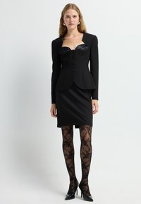 Blazer nero strutturato con collo in satin, sei bottoni, abbinato a una gonna nera aderente e collant neri a fantasia, completato da tacchi alti.