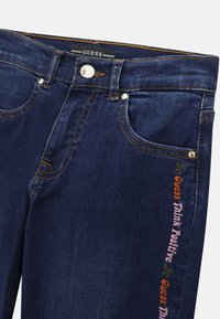 Unos pantalones de mezclilla azul oscuro con costuras naranjas, un botón de metal y bordado lateral que dice "be Guess Think Positive" en varios colores.