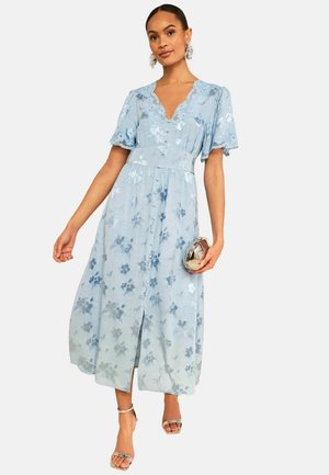 Frau trägt ein hellblaues florales Midikleid mit Spitzenbesatz, flatternden Ärmeln, geknöpfter Taille, hält eine goldene Clutch und trägt silberne Absätze.