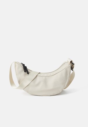 Borsa a tracolla a forma di mezzaluna beige con tracolla in tessuto regolabile e tirazip neri su sfondo bianco.