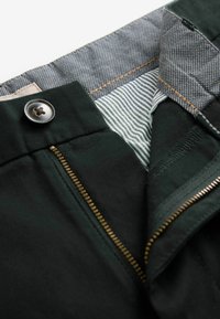Gros plan sur un pantalon foncé avec une fermeture éclair en laiton ouverte, un bouton gris et une doublure en tissu rayé à l'intérieur de la ceinture.