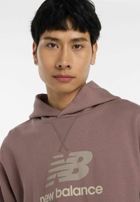 Sudadera de color malva claro hecha de tejido suave, con capucha ajustable y un gran logo en gris contrastante de "New Balance" en la parte frontal.