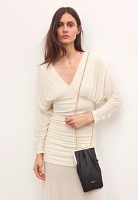 Juodas odinis rankinukas su auksine grandinėlę, turintis struktūrinę rėmą ir tekstūruotą kūną. Modelis vilki šviesiai kreminę suknelę.