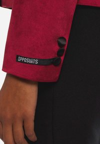 Punainen mokkanahkainen bleiserin hiha, jossa on kolme mustaa nappia ja kudottu "OPPOSUITS" -label. Kankaalla on sileä pinta.