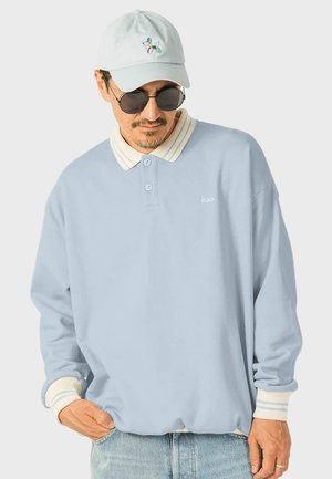 Lyseblå langærmet polo med hvid og lyseblå stribet krave. Udstyret med to knapper og lille broderet logo på brystet.