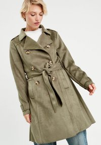 Trench coat de color verde oliva con un diseño de doble botonadura, cintura ceñida con cinturón y mangas largas con puños abotonados. Tecido suave y con textura.